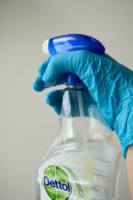 A person wearing light green rubber gloves is holding a stack of white paper or brochures, positioned near a grey wall in an indoor setting. The background shows a shelf or ledge with various cleaning