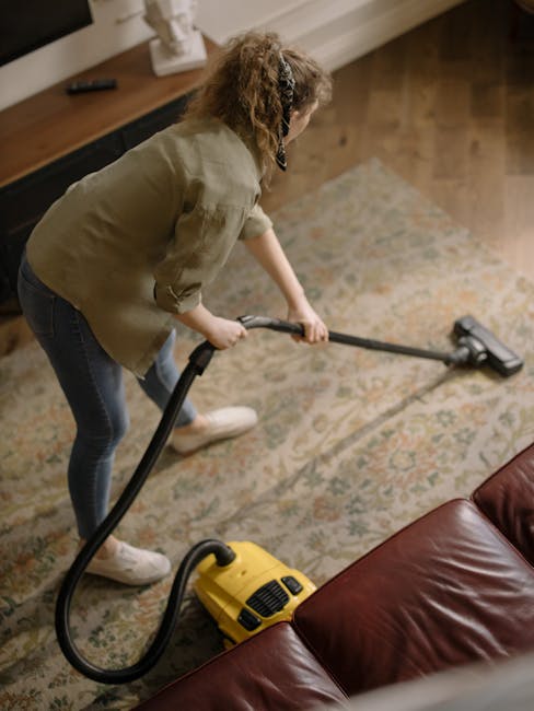 A woman with brown hair tied back, wearing a light blue striped shirt and jeans, is kneeling on a patterned rug and actively cleaning a wooden floor with a blue cloth. She is in a modern living room w