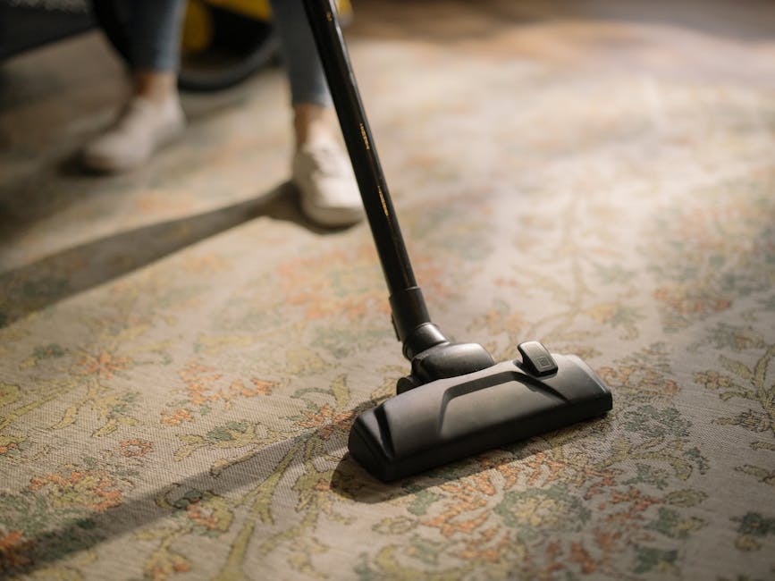 Close-up view of a person vacuuming a traditional patterned area rug with a handheld vacuum cleaner, with focus on the vacuum head and part of the vacuum stick. The rug features intricate floral designs in muted tones of beige, green, and orange, and the room appears well-lit, highlighting the surface's cleanliness and the cleaning process. The person's legs, dressed in casual clothing and sneakers, are visible in the background, standing on the rug. This scene exemplifies routine surface cleaning and deep cleaning practices as part of home hygiene maintenance, with professional cleaning services like Kennington Carpet Cleaners ensuring optimal sanitisation of domestic carpets.
