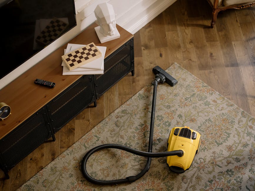 A yellow vacuum cleaner with a black hose and metal wand is positioned on a patterned area rug in a living room. Adjacent to the rug is a wooden floor with a dark finish. On top of a low black metal and wood sideboard, there are a closed remote control, a white bust sculpture, a stack of papers, and two wooden chessboards. The room is illuminated with natural light, highlighting the clean and tidy appearance of the carpet and surrounding surfaces. Kennington Carpet Cleaners' logo and branding are associated with the scene, emphasizing professional surface cleaning and deep cleaning services for residential spaces.