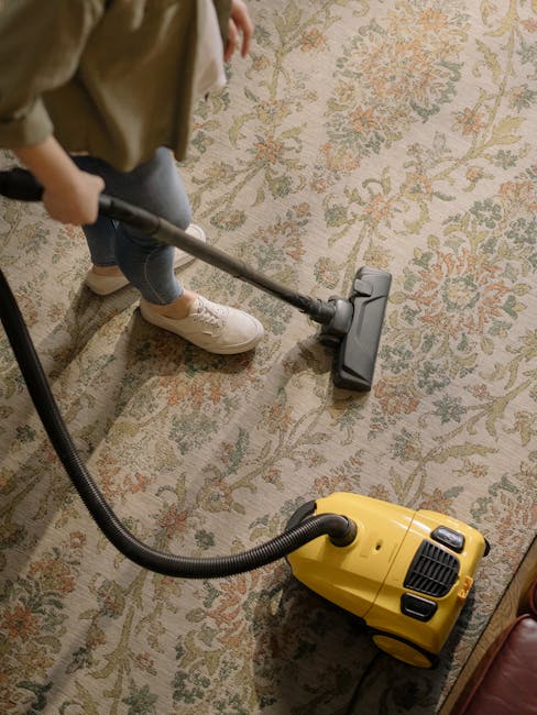 Close-up view of a person vacuuming a traditional patterned area rug with a handheld vacuum cleaner, with focus on the vacuum head and part of the vacuum stick. The rug features intricate floral designs in muted tones of beige, green, and orange, and the room appears well-lit, highlighting the surface's cleanliness and the cleaning process. The person's legs, dressed in casual clothing and sneakers, are visible in the background, standing on the rug. This scene exemplifies routine surface cleaning and deep cleaning practices as part of home hygiene maintenance, with professional cleaning services like Kennington Carpet Cleaners ensuring optimal sanitisation of domestic carpets.