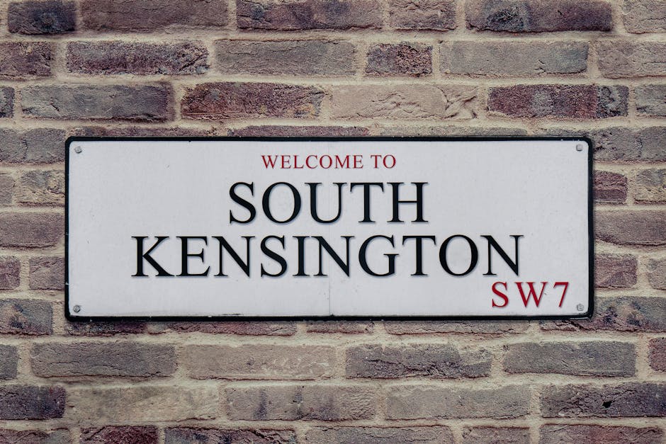 A rectangular white metal street sign mounted on a red and gray brick wall, displaying the text 'WELCOME TO SOUTH KENSINGTON SW7' in black and red lettering. The sign is attached with four screws, one